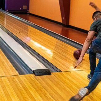 A person bowls at a modern alley with colorful lanes and arcade games in the background. Bright lights illuminate the area.