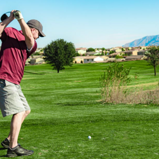 A lush golf course landscape with green fairways, mountains in the distance, and a clear blue sky overhead.