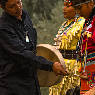 Man in traditional regalia with a feathered headdress stands on a stage, engaging with an audience during a cultural event.