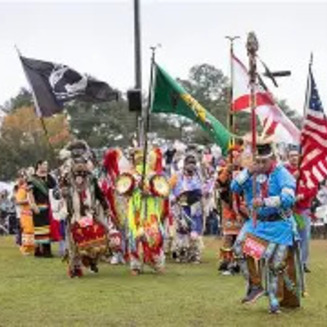 Native dancers in colorful regalia with feathered headdresses perform at Poarch Creek Pow Wow, surrounded by spectators in an outdoor arena.